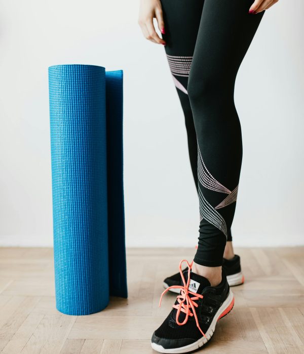 A woman in leggings and sneakers standing with a yoga mat indoors.